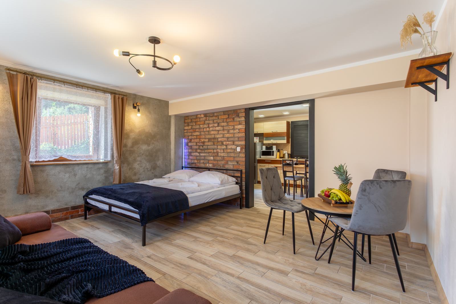 Przestronny pokój w stylu loft z ceglaną ścianą, dwuosobowym łóżkiem i stolikiem, w głębi widoczna kuchnia